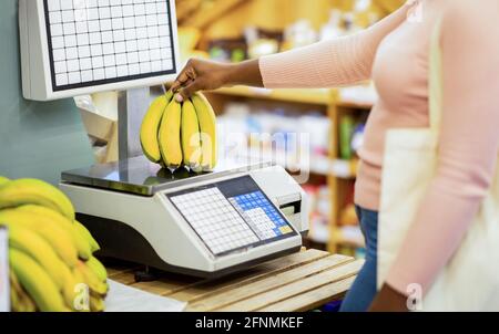 Vista tagliata di giovane donna nera che pesa banane su bilance al supermercato, guardaroba Foto Stock