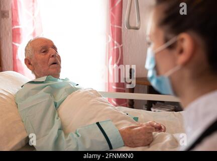 Uomo anziano seduto in letto medico in ospedale e giovane infermiere con maschera protettiva che lo visita Foto Stock