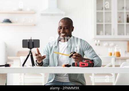 Handyman nero che registra video sulla riparazione e la ristrutturazione, guardando il cellulare in webcam e tenendo il nastro di misurazione, Foto Stock