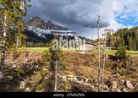 Capanne di legno in un prato ai piedi di cime rocciose innevate nelle Alpi con nuvole di stom che si profilano in una giornata di primavera. Un'insenatura è visibile in primo piano. Foto Stock