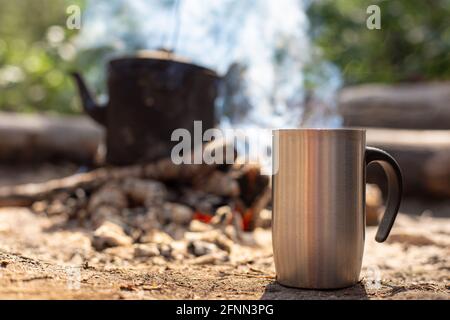 La tazza di metallo con una bevanda calda si trova a terra, sullo sfondo di un falò con un bollitore, in un'escursione, nella foresta. Foto Stock