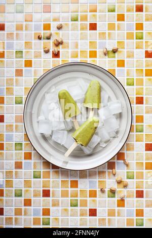 Popsicle di pistacchio in casa surgelato in ciotola di ghiaccio su tavola di tegole a mosaico. Papsicle rinfrescante, succo verde congelato sul bastone. Vista dall'alto, spazio di copia. Foto Stock