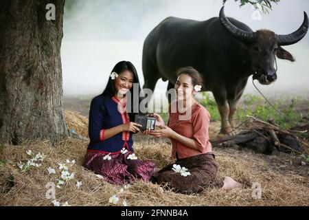 Due donne asiatiche che indossano la cultura tradizionale tailandese di stile esan seduto sul campo, mentre ascoltano radio di stile vintage contro bufali e bac fattoria Foto Stock