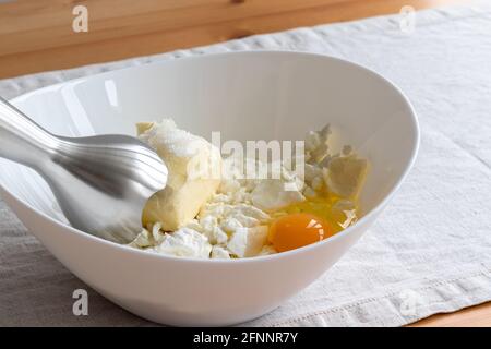 In un recipiente bianco, il formaggio, il burro, le uova e lo zucchero vengono fatti in casa - ingredienti da mescolare con un frullatore a immersione su una tovaglia di lino su un tavolo di legno. Il processo Foto Stock