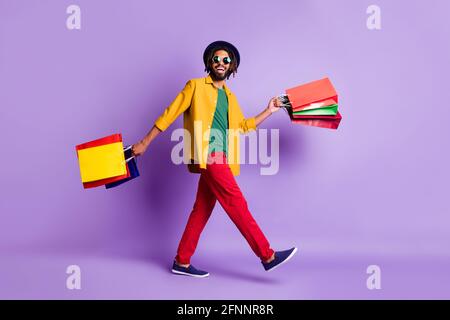 Foto a lunghezza intera di dolce cute ragazzo di pelle scura vestito camicia gialla che cammina tenendo borse isolato colore viola sfondo Foto Stock