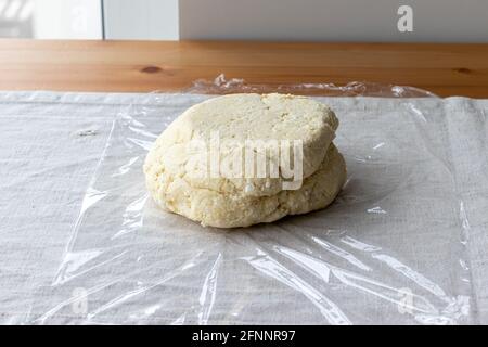 Impasto di cagliata cruda con pezzi di formaggio da cottage su un involucro di plastica su una tovaglia di lino su un tavolo di legno. Il processo di preparazione di bagel o croissant cagliati Foto Stock