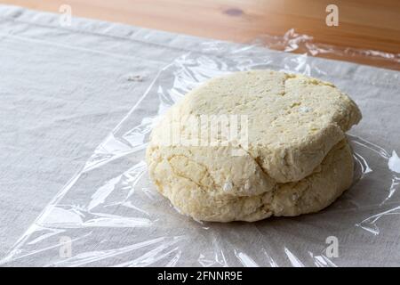 Impasto di cagliata cruda con pezzi di formaggio da cottage su un involucro di plastica su una tovaglia di lino su un tavolo di legno. Il processo di preparazione di bagel o croissant cagliati. Foto Stock