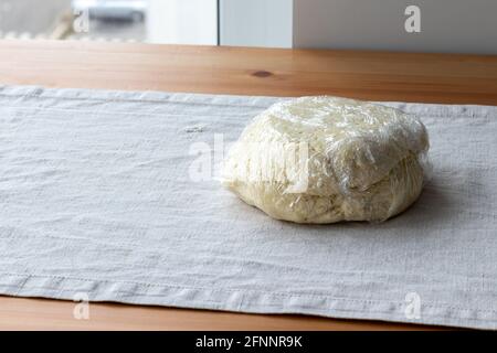 L'impasto di cagliata cruda con le punte di formaggio di cottage è avvolto in un involucro di plastica su una tovaglia di lino su un tavolo di legno. Il processo di fabbricazione dei bagels cagliati o cr Foto Stock