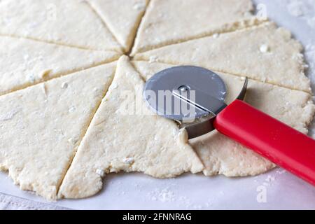 Pasta arrotolata di cagliata cruda con punte di formaggio di cottage tagliate in otto triangoli con un coltello rosso rotondo per pizza su carta da forno closeup. Il processo di Foto Stock