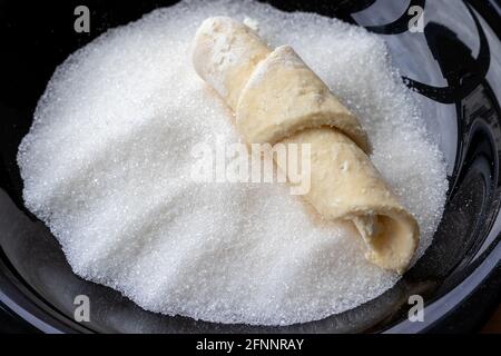 Una busta, attorcigliata da pasta cruda di formaggio di cottage, giace su un piatto nero con lo zucchero per rotolare in esso. Il processo di preparazione di bagel o croissant cagliati Foto Stock