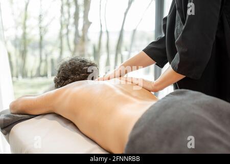 Massaggiatore femminile che fa massaggio al cliente maschile nel complesso termale. Benessere e tempo libero. Mettere a fuoco su una schiena con le mani. Primo piano. Foto Stock