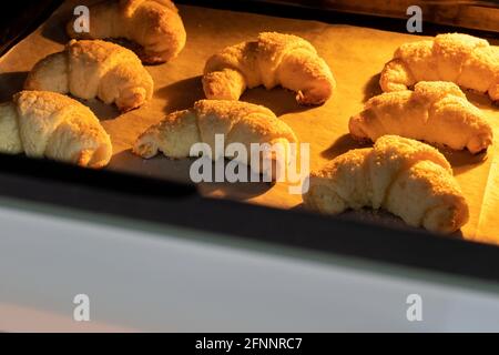 Croissant ghiacciati al forno su una teglia ricoperta di carta pergamena. Bagel dolci da un impasto casolare di formaggio cotto in un forno ajar caldo. Foto Stock