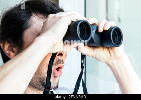 Bell'uomo con un volto sorpreso guardando attraverso binocoli alla finestra con una vista della casa vicina per osservare i vicini, l'ambiente Foto Stock