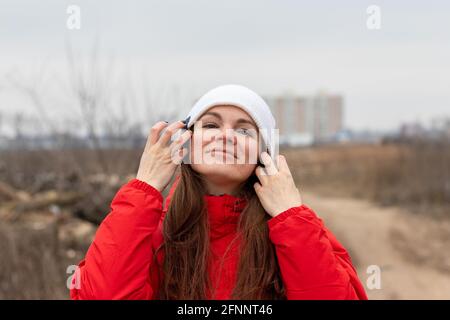 Felice bella giovane donna con capelli lunghi in maglia bianca cappello e giacca rossa rimuove i capelli dal viso con le sue mani sulla strada su un aut nuvoloso Foto Stock