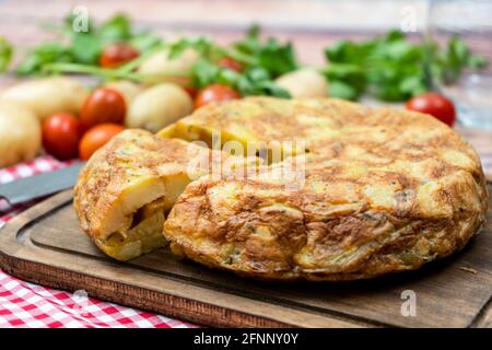 Vista normale di una tipica frittata spagnola di patate o patate con una porzione separata con patate e pomodori ciliegie in un ambiente rustico. Tradizionale Foto Stock