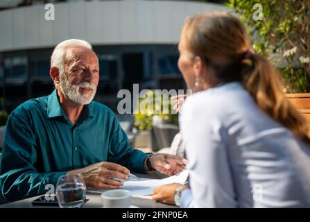 Giovane donna graziosa di affari e uomo maturo che firma l'accordo dentro caffetteria all'aperto Foto Stock