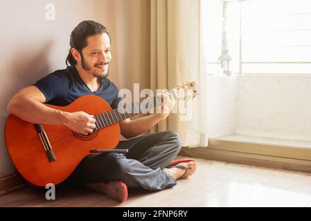 Giovane latino seduto sul pavimento che suona la chitarra a. casa in soggiorno Foto Stock