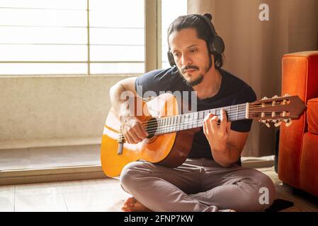 Giovane latino con le cuffie seduto sul pavimento che gioca chitarra a casa in soggiorno Foto Stock
