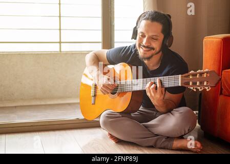 Giovane latino con le cuffie seduto sul pavimento che gioca chitarra a casa in soggiorno Foto Stock