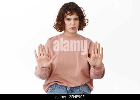 Stop, indietro. Grave ragazza riccia, mostrando blocco proibire gesto, rifiutare qualcosa, stare lontano segno, dire a distanza sociale, in piedi Foto Stock