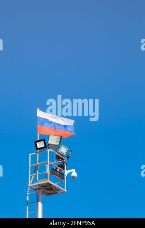 Illuminazione della parte superiore dell'albero con bandiera russa, fari e telecamera CCTV. Primo piano contro il cielo blu chiaro. È presente uno spazio per la copia. La tela della bandiera si sviluppa uniformemente nel vento. Foto Stock