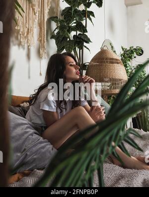 L'attraente giovane donna si rilassa su un comodo letto in una camera da letto accogliente. Interni boho chic con piante di casa e macrame Foto Stock