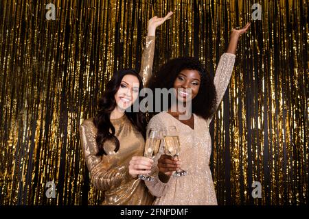 Foto di due belle fidanzate tenere bicchieri spumante sollevare le mani indossano un abito lucido con fondo in tinsel isolato Foto Stock