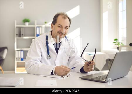 Un medico di sesso maschile amichevole si siede nel suo ufficio di fronte a un computer portatile e compila le cartelle cliniche. Foto Stock