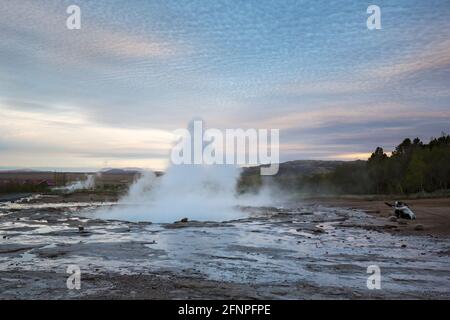 Due giovani donne si siedono accanto al geyser in eruzione scattando foto Sui loro telefoni all'alba nell'Islanda meridionale Foto Stock