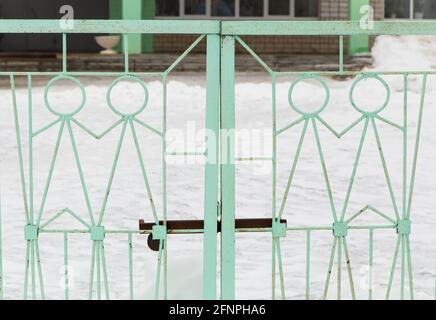 Cancelli in ferro con figure metalliche di persone di colore verde chiaro. Un traliccio fatto di vergelle metalliche con un chiavistello contro lo sfondo dell'edificio scolastico fuori fuoco. Caldo giorno d'inverno, la neve si trova. Foto Stock