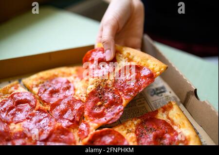 Mano di donne con le unghie graziose che afferrano la fetta della pizza dalla scatola del take away. Peperoni alla pizza con erbe e formaggio e souce. Foto Stock