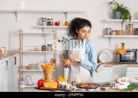 Giovane donna con mortaio e pestello utilizzando il computer portatile in cucina Foto Stock