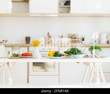 Cucina cibo sano. Pasta italiana, verdure fresche, semi di zucca, scaglie e altri ingredienti su tavolo di legno bianco contro sfondo offuscato da cucina in moderno appartamento luminoso Foto Stock