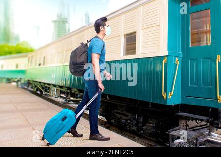 Uomo asiatico in maschera con zaino e valigia sulla stazione ferroviaria. Viaggiare nel nuovo normale Foto Stock