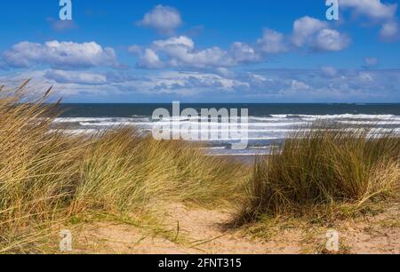 Un'allettante vetta attraverso dune di sabbia verso un cielo blu brillante e il mare con onde infrangenti. Presa su Bambburgh Beach in Northumberland Foto Stock