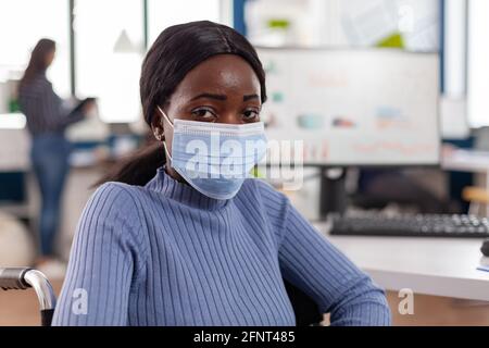 Nero project manager con disabilità disabili maschera paralizzata indossabile guardando la fotocamera seduta immobilizzata in sedia a rotelle, nuovo normale ufficio di lavoro al progetto finanziario. Foto Stock