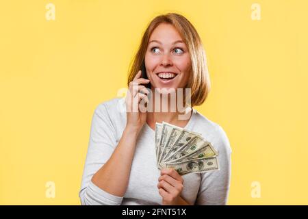 Una donna felice sorprende tiene un ventilatore dei dollari e parla al telefono. Sfondo giallo. Il concetto di sconti e vendite. Foto Stock