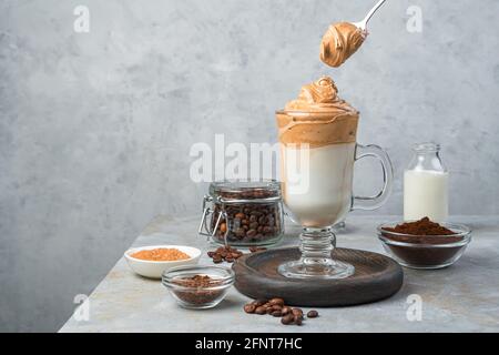 Caffè Dalgona in un bicchiere trasparente e un cucchiaio con schiuma su fondo grigio. Vista orizzontale, spazio di copia. Foto Stock