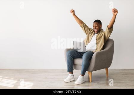 Joyful African Male Using laptop shaking Fists over Grey Wall Foto Stock