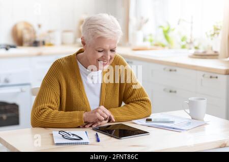 Gioiosa donna anziana dai capelli grigi che usa il tablet digitale in cucina Foto Stock