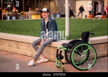 un giovane disabile si siede accanto a una sedia a rotelle e le mostra il dito medio Foto Stock