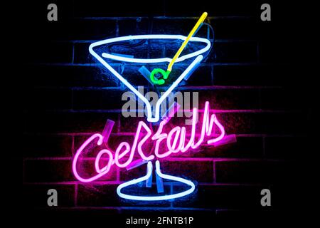 COCKTAIL con iscrizione al neon viola brillante e bicchiere da cocktail blu stilizzato con cannuccia gialla su fondo muro di mattoni scuri. Immagine vintage con toni scuri. Foto Stock