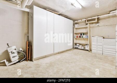 Vari ripiani di stoccaggio e armadio situati sul pavimento in shabby vicino tubi e porta in cantina Foto Stock