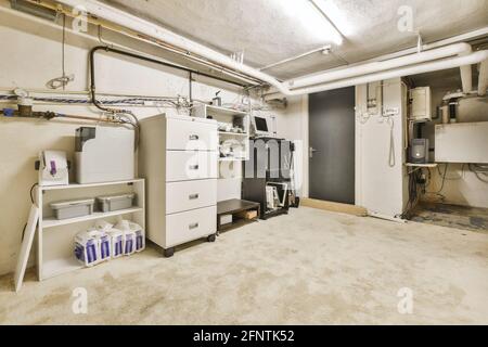 Vari ripiani di stoccaggio e armadio situati sul pavimento in shabby vicino tubi e porta in cantina Foto Stock