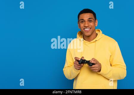 KIEV, UCRAINA - 22 MARZO 2021: Sorridente afroamericano che tiene il joystick in mani isolate su blu Foto Stock