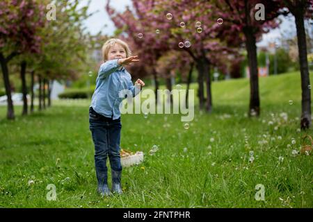 Bambino biondo, ragazzo carino in abbigliamento casual, giocando con bolle di sapone nel parco, correre felicemente Foto Stock