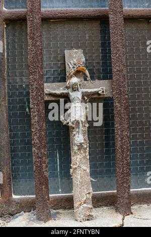 Una croce di Cristo coperta di vecchi ciottoli è incuneata tra due montanti in metallo arrugginito di fronte ad un vecchio finestra in vetro Foto Stock