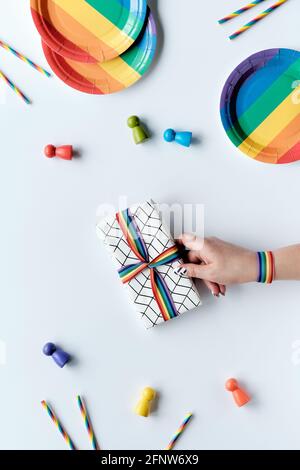 LGBTQ comunità orgoglio mese rainbow concetto piatto laici. La mano con il nastro arcobaleno contiene il regalo confezionato. Piatti da festa arcobaleno, paillette di carta da bere. Disposizione piatta Foto Stock