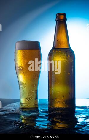 bottiglia di birra versata in acqua su una bottiglia blu di birra con un bicchiere di birra schiumosa su sfondo blu, sull'acqua Foto Stock