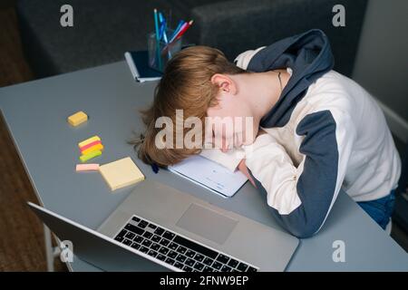 Primo piano del ragazzo stanco che dormiva sul libro degli esercizi di fronte Di laptop dopo la lezione online via Internet Foto Stock